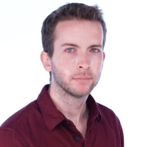 A profile photo of Los Angeles Culture Index reviewer/critic Ethan James Hulbert, wearing a soft red button-up shirt with open collar, featuring short brown hair and a gaze looking forward at the viewer, against a plain white background.