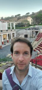 Man with short brown hair and striped shirt taking a selfie in the Getty Villa ancient theater with red seats and hillside background