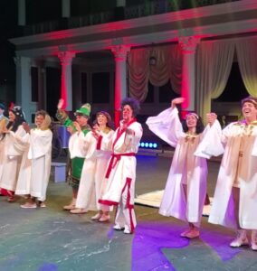 Actors in white robes and colorful costumes hold hands and bow on stage with red-lit columns and draped curtains in the background