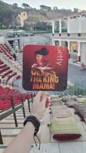 Hand holding a fan with caricature of Elvis and text Oedipus The King Mama at Getty Villa outdoor theater with red and beige seats