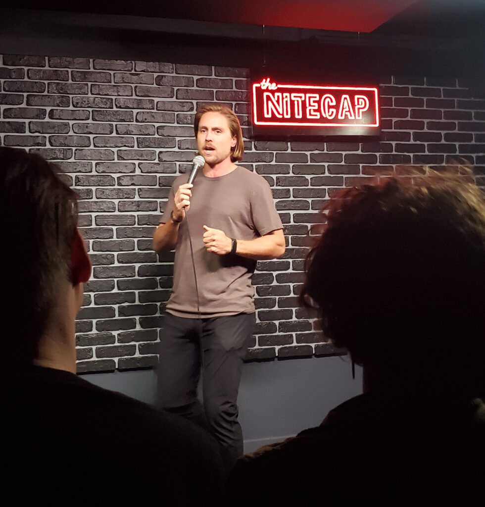 a man at the nitecap comedy club with black and white brick wall and neon sign behind him
