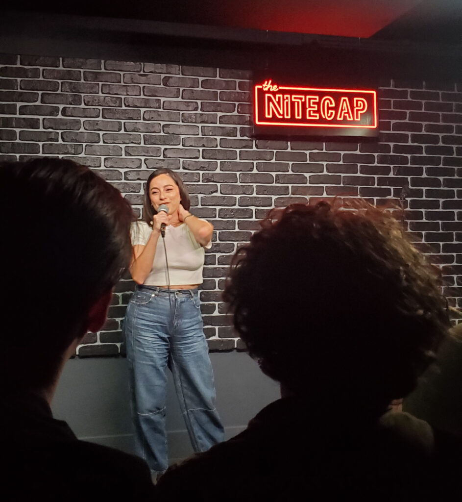jess alaniz comedian - a woman doing stand-up comedy in front of a brick wall