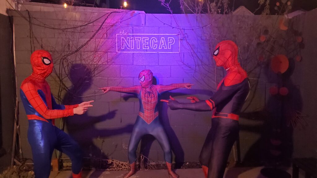 three people in different spider-man costumes all pointing at one another, brick wall behind that says the nitecap